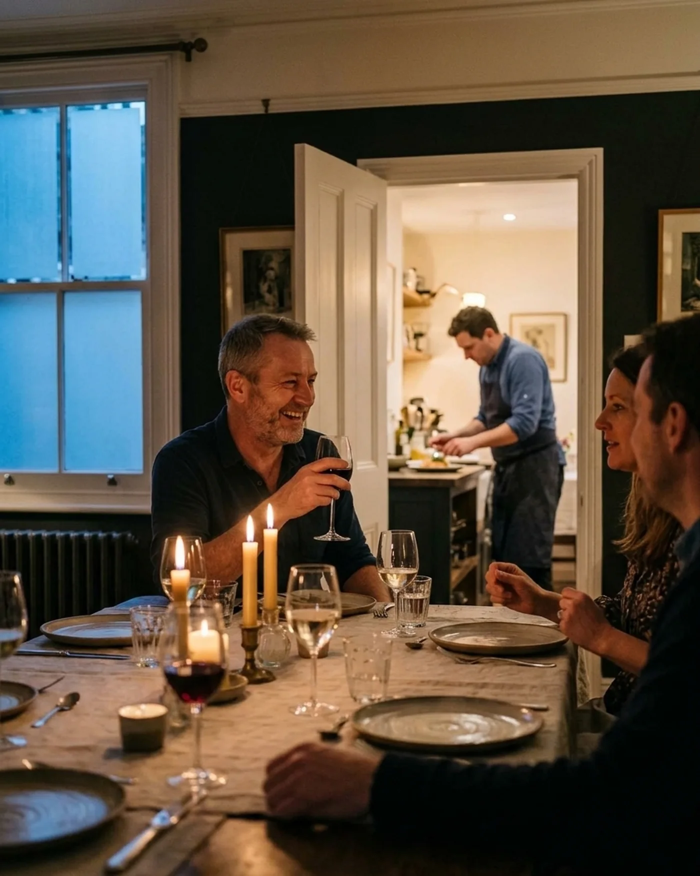 A host laughing with guests at a candlelit table in a London dining room while the chef plates the next course through the kitchen doorway